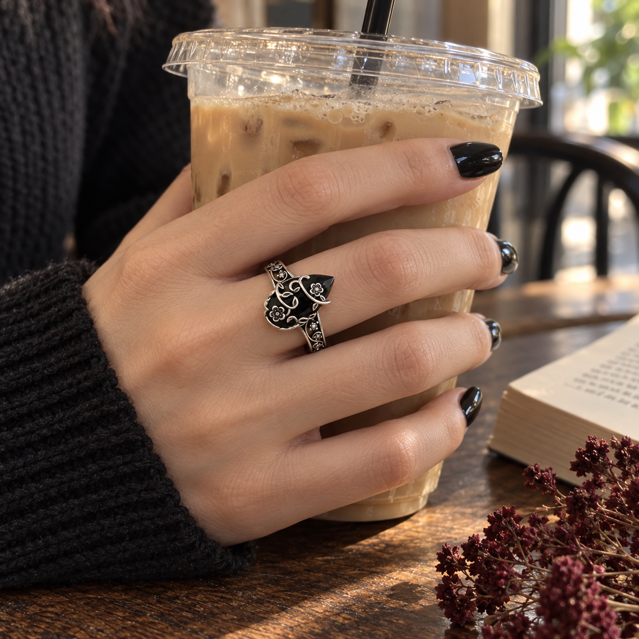 Black-manicured hand wearing the floral ring while holding an iced coffee