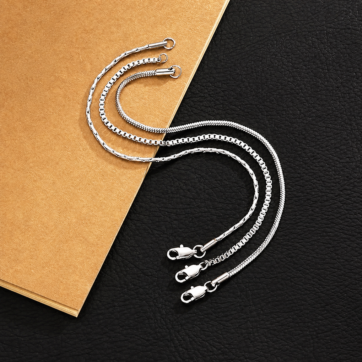 Veyron box, snake, and patterned herringbone-style bracelet chains arranged on a blank tan paper and dark surface